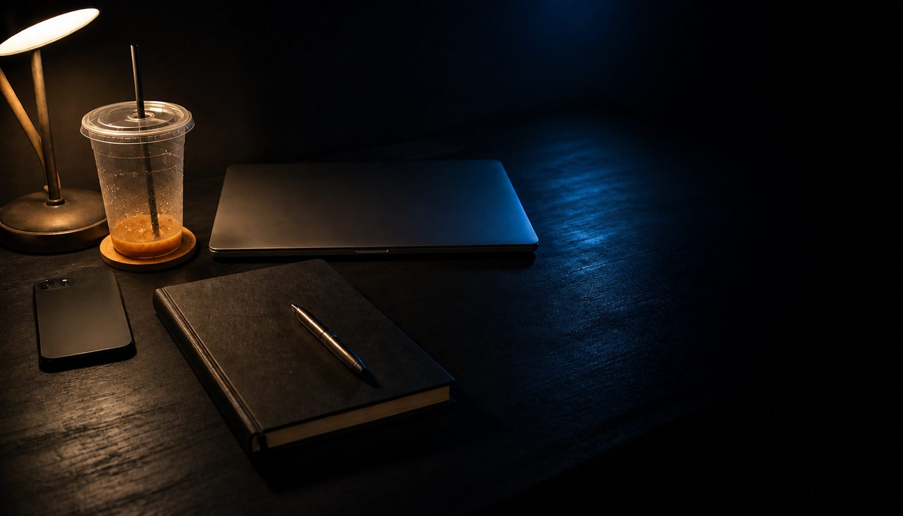 A closed notebook with a pen resting on top and a closed laptop on a dark desk, warm lamp light from the left and a cool blue spill from the right