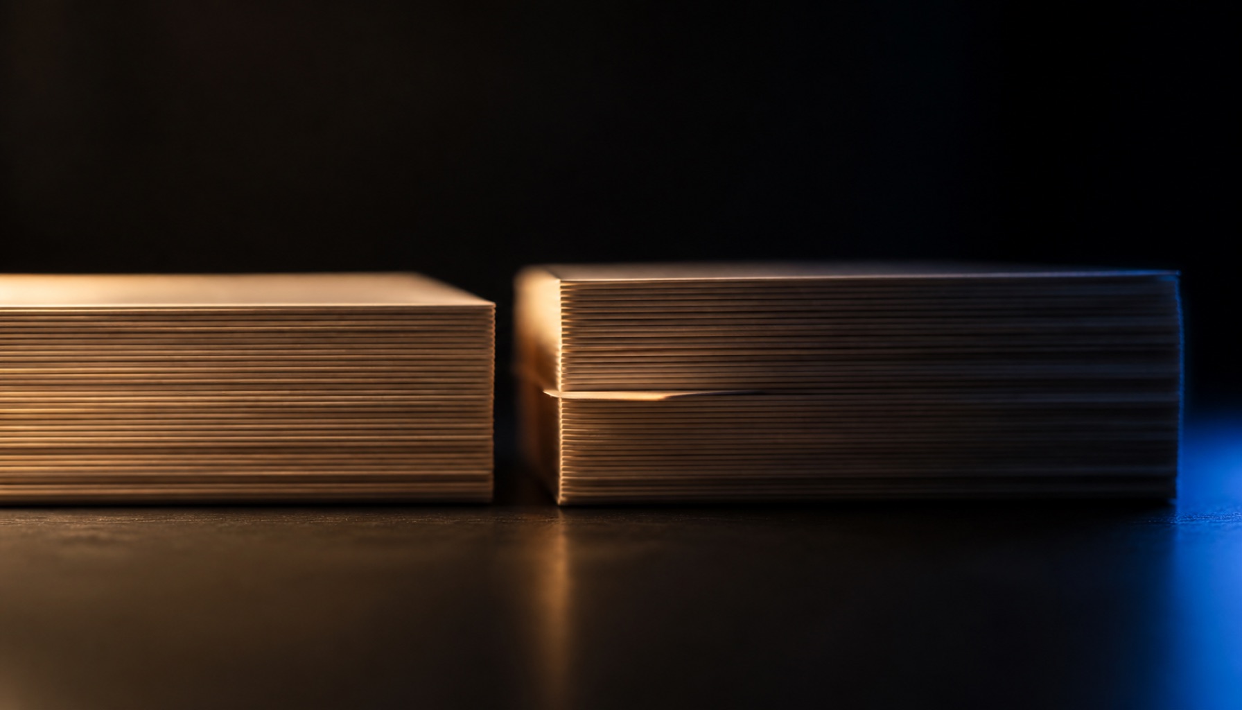 Two nearly identical stacks of paper seen edge-on across a dark desk under a warm lamp, the right-hand stack slightly taller with one sheet protruding out of line